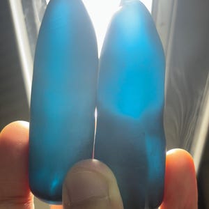 Puede incluir: Dos piedras azules translúcidas, lisas y alargadas con extremos redondeados. Las piedras se sostienen frente a una fuente de luz brillante, lo que ilumina su color azul profundo y revela sutiles variaciones internas y un aspecto suave.