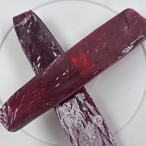 Peut inclure: Deux barres rectangulaires allongées en matériau rouge rubis profond et translucide, disposées en forme de X. Les surfaces présentent un aspect cristallin texturé avec des fractures internes visibles et des reflets lumineux sur la couleur.