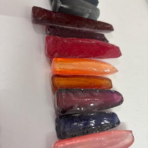 May include: A collection of ten elongated, wedge-shaped resin pieces arranged in a vertical column. The pieces feature a variety of translucent and opaque colours, including light blue, dark grey, deep red, bright orange, purple, and light pink.