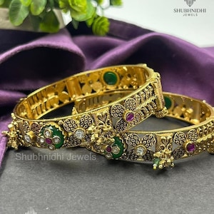 May include: Two gold bangles with detailed floral and geometric patterns. The bangles are adorned with green, red, and clear gemstones. The text "Shubhnidhi Jewels" is visible on the bangles.