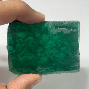 Puede incluir: Un espécimen mineral rectangular de color verde translúcido con una textura cristalina rugosa. La piedra muestra tonos esmeralda profundos con parches más claros y nublados, además de bordes irregulares y sin pulir.