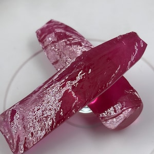 Puede incluir: Dos fragmentos de cristal alargados, translúcidos y de color rosa intenso, con una superficie rugosa y texturizada y bordes afilados y angulosos. Los cristales están colocados en forma de cruz, mostrando un tono magenta vibrante y una calidad reflectante.