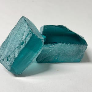 Può includere: Due pezzi di vetro traslucido color verde acqua con bordi ruvidi e frastagliati e una superficie superiore piatta e liscia. I pezzi di vetro hanno un colore blu-verde profondo e saturo e presentano una consistenza cristallina.