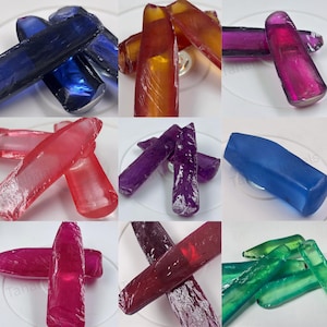 Puede incluir: Una colección de nueve paneles que muestran varias barras alargadas, translúcidas y pulidas de vidrio o piedras preciosas en colores vibrantes, incluyendo azul profundo, ámbar, magenta, rojo brillante, morado, azul mate, rosa, granate y verde esmeralda.