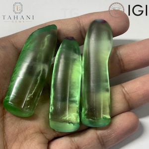 Puede incluir: Tres piedras preciosas verdes translúcidas y alargadas con superficies lisas y redondeadas, con puntas de tonos púrpuras sutiles. Las piedras presentan un acabado pulido que capta la luz y están dispuestas una al lado de la otra.