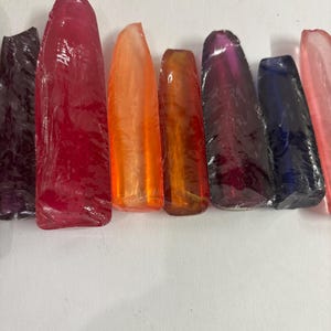 May include: A collection of seven elongated, translucent glass shards arranged in a row. The pieces display a vibrant spectrum of colours including deep purple, bright red, orange, amber, magenta, dark blue, and a pale pink, each with a smooth finish.