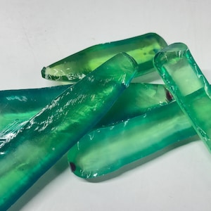 May include: A collection of translucent, elongated, and irregularly shaped green glass shards. Each piece features a smooth, polished surface with varying gradients of emerald and lime green hues, showcasing a glossy finish and sharp edges.