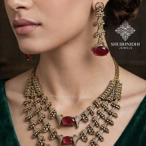 Puede incluir: Un conjunto de collar y pendientes dorados con grandes piedras rojas ovaladas. El collar tiene tres capas con detalles intrincados y pequeñas piedras transparentes. Los pendientes tienen un diseño similar. La marca "SHUBHNIDHI JEWELS" es visible.