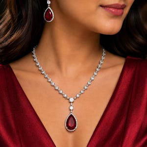 May include: A dazzling jewelry set featuring a necklace and matching earrings. The necklace and earrings showcase a teardrop-shaped, deep red gemstone, surrounded by a halo of sparkling white stones. The jewelry is set against a rich, red background.