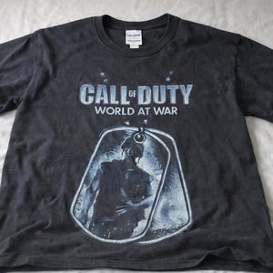 May include: Black t-shirt featuring the text "CALL OF DUTY WORLD AT WAR" in blue and white. A dog tag graphic with a soldier is below the text. The shirt is made of a soft material.