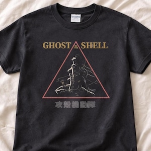 May include: A black short-sleeve t-shirt featuring a graphic print of a figure inside a red triangle. The text GHOST IN THE SHELL is printed in gold above the triangle, and Japanese characters are printed in white below the triangle.