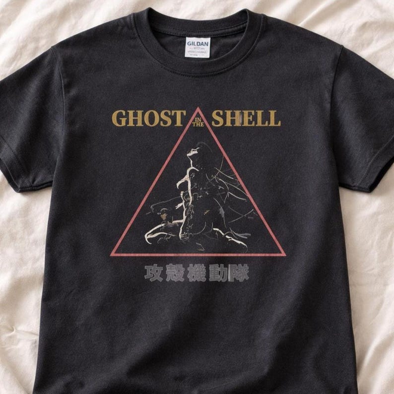 May include: A black short-sleeve t-shirt featuring a graphic print of a figure inside a red triangle. The text GHOST IN THE SHELL is printed in gold above the triangle, and Japanese characters are printed in white below the triangle.