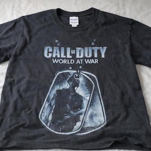 May include: A black short-sleeved t-shirt featuring the Call of Duty World at War logo in white and blue text. Below the text is a graphic of a soldier silhouette inside a dog tag shape, with a Gildan brand tag at the inner neckline.