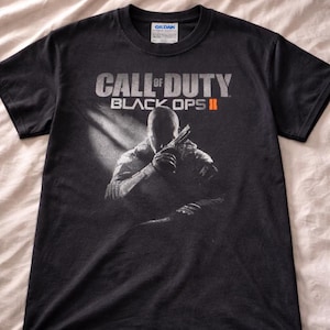 May include: A black short-sleeve t-shirt featuring the Call of Duty Black Ops II logo in white and orange text. Below the logo is a graphic of a soldier in silhouette holding a firearm, with a light beam effect extending from the upper left side.
