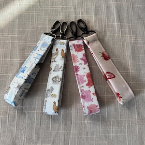 May include: Four fabric keychains with black metal clasps. The keychains feature various patterns: blue dinosaurs, chickens and eggs, pink pigs, and strawberries on a pink background. The keychains are made of fabric.