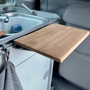 Puede incluir: Una configuración de cocina compacta para furgoneta camper con fregadero de acero inoxidable, tablas de cortar de madera y una toalla colgando de un riel. Las tablas de cortar son de madera clara. El interior es gris claro.