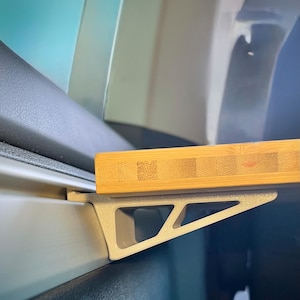 Può includere: Una staffa triangolare grigio chiaro per accessori auto con un vassoio in bambù. La staffa è progettata per essere fissata alle finiture interne di un'auto. Il vassoio in bambù è rettangolare e presenta venature naturali.