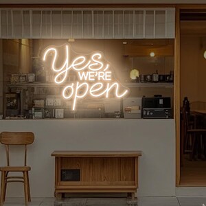 Op de afbeelding: Een neonbord in een etalage leest "Yes, We're Open" in een cursief lettertype. Het bord gloeit met een warme, uitnodigende gloed. Een houten stoel en kast staan buiten de winkel.