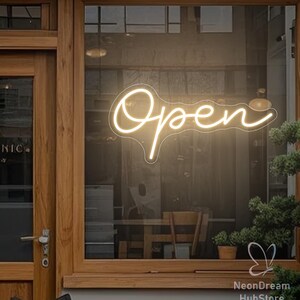 May include: A glowing neon sign in a shop window displays the word "Open" in a cursive font. The sign is a warm, off-white color and is mounted on a glass window. The shop's wooden door and interior are visible.