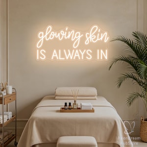 May include: A wall-mounted neon sign featuring the text glowing skin IS ALWAYS IN in a warm white light. The sign is mounted above a treatment bed covered in a beige cloth, with a wooden tray holding small bottles and folded white towels.