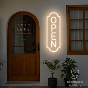 Könnte beinhalten: Ein leuchtendes Neon-Schild mit dem Wort "OPEN" in vertikaler Ausrichtung ist an einer weißen Wand angebracht. Eine Holztür und ein Fenster rahmen die Szene ein, mit Topfpflanzen darunter. Das Schild beleuchtet den Laden.