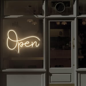 Op de afbeelding: Een oplichtend neonbord in een etalage toont het woord "Open" in een sierlijk lettertype. Het bord heeft een warme, uitnodigende gele kleur, die contrasteert met het donkere interieur dat door het glas zichtbaar is.