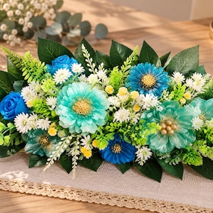 Può includere: Un centrotavola floreale composto da un mix di fiori artificiali blu e turchesi, tra cui rose e margherite, arricchito da piccoli fiori bianchi, boccioli gialli e varie foglie verdi simili a felci, disposti in una lunga composizione orizzontale.