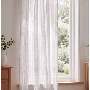 May include: White sheer curtain panel with vertical patterned stripes. The curtain hangs in front of a window, allowing natural light to filter through. The curtain is made of a lightweight fabric and is likely used for privacy and decoration.