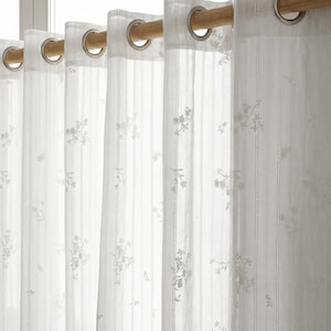 May include: Sheer white curtains featuring a delicate vertical striped pattern and small embroidered floral motifs. The fabric is suspended from a light-toned wooden rod by evenly spaced silver-colored metal grommets along the top header.