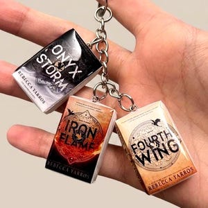 Peut inclure: Un porte-clés en métal avec trois breloques en forme de livres miniatures. Les breloques présentent les couvertures de Onyx Storm, Iron Flame et Fourth Wing de Rebecca Yarros, avec des tons noir, orange et crème, fixées à un anneau argenté.