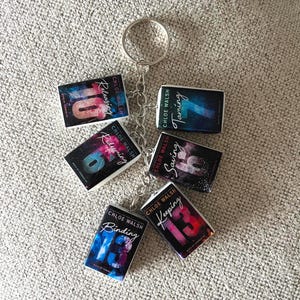 Può includere: Un portachiavi in metallo argentato con sei ciondoli rettangolari a forma di libro in miniatura. Ogni ciondolo mostra una copertina di un libro di Chloe Walsh con i titoli Releasing 10, Taming 7, Redeeming 6, Saving 6, Binding 13 e Keeping 13.
