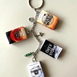 May include: A silver-toned metal keyring featuring four miniature book charms titled Fourth Wing, Iron Flame, Onyx Storm, and Threshing Day by Rebecca Yarros, accented with a small spiral charm, a star charm, and green beads.