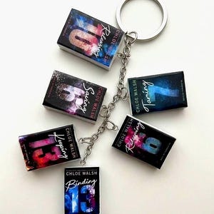May include: A silver metal keyring featuring six miniature book charms. Each charm displays a book cover by Chloe Walsh with titles including Releasing 10, Saving 6, Taming 7, Redeeming 6, Keeping 13, and Binding 13, all with colourful, nebula-like graphic designs.