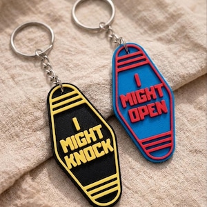 Könnte beinhalten: Zwei Schlüsselanhänger mit silbernen Ringen. Einer ist schwarz mit gelbem Text "I MIGHT KNOCK". Der andere ist blau mit rotem Text "I MIGHT OPEN". Beide haben ein Retro-Motel-Schlüssel-Design.