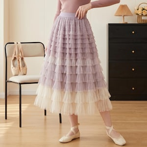 Può includere: Una gonna a strati in tulle nei toni del lavanda e crema. La gonna presenta più strati di tessuto morbido e fluido, creando una silhouette voluminosa. La modella indossa ballerine. Un paio di scarpette da ballo poggia su una sedia.