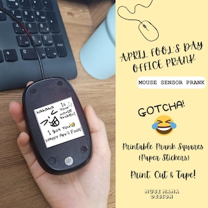May include: A black computer mouse with a white sticker on the bottom, featuring text and a cartoon face. The sticker reads "Is your mouse broken? I got you! Happy April Fools!" and is part of an office prank.