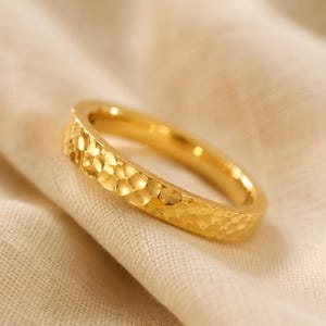 Puede incluir: Un anillo de oro con textura martillada. El anillo es de color oro brillante y tiene una superficie texturizada. El anillo está sobre una tela color crema.