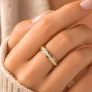May include: A gold band ring with a row of small, clear stones set between two gold bands. The ring is on a person's finger, and the background is a soft, neutral color.