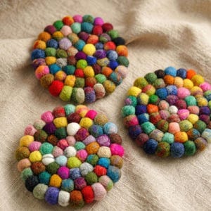 Puede incluir: Tres posavasos redondos de fieltro colorido. Cada posavasos está hecho de pequeñas bolas de fieltro muy juntas en una variedad de colores, incluyendo rojo, amarillo, verde, azul y rosa. Los posavasos están colocados sobre una tela de color neutro.