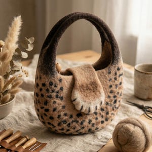 May include: A handmade felt handbag with a unique leopard print pattern. The bag features a rounded shape with a dark brown handle and a flap closure. The bag's body is tan with black spots, and a small wooden bead is attached to the handle.