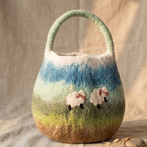 May include: A felted wool bag with a rounded shape and a green handle. The bag features a landscape design with blue, green, and brown hues, and two white sheep with pink accents.