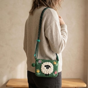 K&ouml;nnte beinhalten: Gr&uuml;ne Filz-Umh&auml;ngetasche mit Schafmotiv. Die Tasche zeigt ein wei&szlig;es Schaf mit schwarzem Gesicht und K&ouml;rper, umgeben von bunten Blumen. Der Riemen ist gr&uuml;n und verstellbar.