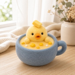 May include: A needle-felted yellow chick figurine sits inside a blue felted mug filled with yellow felted soup. The chick has black bead eyes, an orange beak, and a small yellow square on its head. Small yellow and white felted spheres float in the soup.