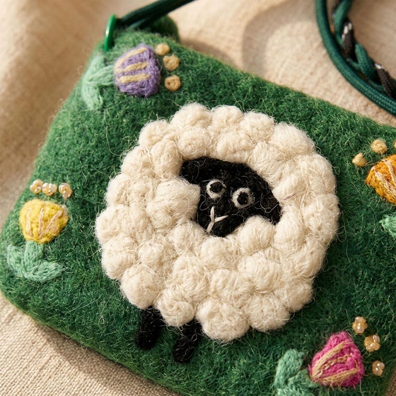 K&ouml;nnte beinhalten: Eine gr&uuml;ne Filztasche mit einem wei&szlig;en Schafdesign. Das Schaf hat ein schwarzes Gesicht und Beine mit einem flauschigen wei&szlig;en Wollk&ouml;rper. Die Tasche ist mit gestickten Blumen in Gelb, Lila und Rosa verziert. Ein gr&uuml;ner Riemen ist an der Tasche befestigt.
