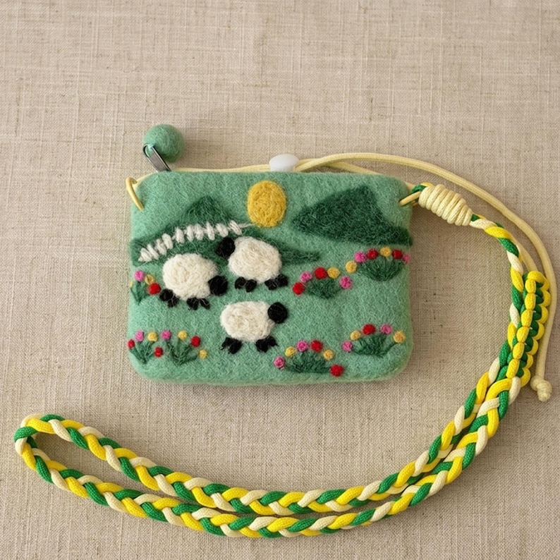 K&ouml;nnte beinhalten: Eine gr&uuml;ne Filztasche mit einem Landschaftsdesign mit drei Schafen, Bergen und Blumen. Die Tasche hat einen gelben geflochtenen Riemen und einen kleinen gr&uuml;nen Kugelrei&szlig;verschluss. Misst etwa 10 cm breit und 7,6 cm hoch.