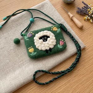 K&ouml;nnte beinhalten: Eine gr&uuml;ne Filz-Umh&auml;ngetasche mit einem wei&szlig;en Schafdesign und schwarzen Details. Die Tasche hat einen geflochtenen gr&uuml;nen Riemen und bunte Blumenmuster. Die Tasche ist etwa 13 cm breit und 8 cm hoch.