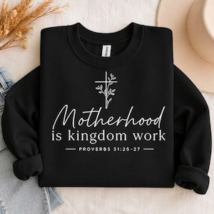Könnte beinhalten: Schwarzes Sweatshirt mit dem weißen Text "Motherhood is kingdom work". Ein weißes Kreuz mit Blättern befindet sich über dem Text, und der Bibelvers Sprüche 31:25-27 steht darunter. Das Sweatshirt ist gefaltet.