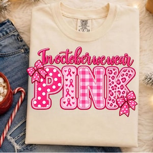 Op de afbeelding: Een crèmekleurig t-shirt met de tekst In October We Wear Pink in een vetgedrukt magenta lettertype. Het woord PINK is versierd met witte stippen, roze linten, een roze gingham ruitpatroon en een roze luipaardprint met twee strikken.