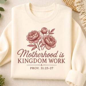 Puede incluir: Sudadera de color crema con un diseño floral y el texto "Motherhood is KINGDOM WORK" con "PROV. 31:25-27" debajo. El diseño floral presenta tres peonías rosas con hojas verdes. La sudadera tiene cuello redondo y mangas largas.