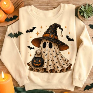May include: A cream-colored sweatshirt featuring a graphic of a ghost wearing a large black witch hat with a buckle. The ghost is covered in a pattern of small black bats and stars, and it is holding a black jack-o'-lantern bucket. Several black bats and orange star-shaped sparkles surround the central ghost figure.